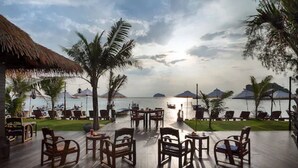 An outdoor seating area with wooden tables and chairs, overlooking a body of water with boats and a thatched-roof structure. - Mali Resort Sunrise Beach (Koh Lipe)