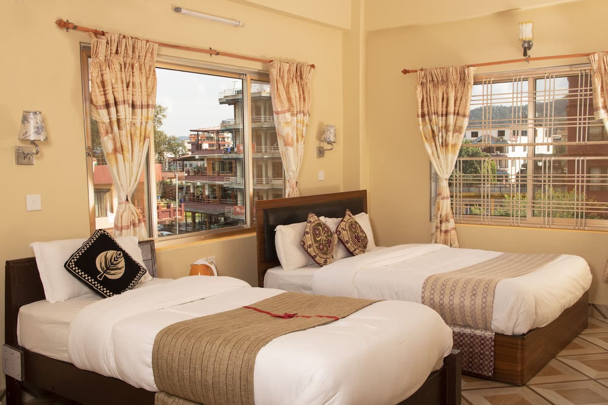 A hotel room with two beds, a window with curtains, a lamp, and a view of buildings outside.