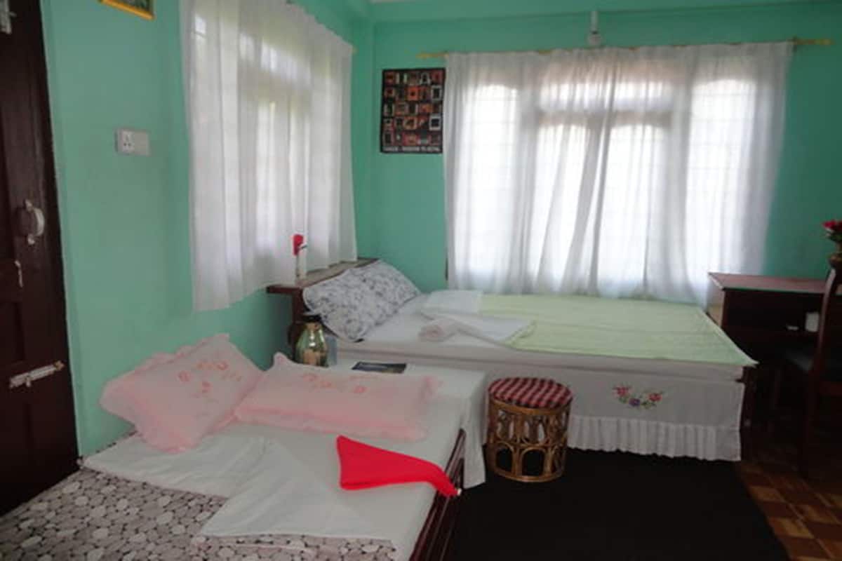A bedroom with a bed, a nightstand, a small table, a wicker stool, and a window with curtains.