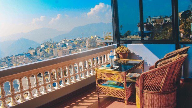 Un balcón con mobiliario de mimbre, una mesa con una planta y vistas a la ciudad y a las montañas.