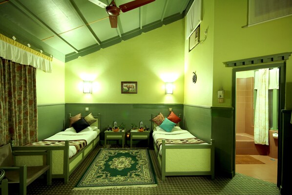 Una habitación con dos camas individuales, una mesita pequeña y una silla. Hay dos mesitas de noche con lámparas. La pared es verde y tiene un ventilador de techo.