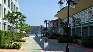 A hotel corridor with a view of the sea, featuring a tiled walkway, lamp posts, and greenery.