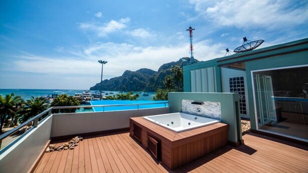 A wooden deck with a hot tub and a glass-enclosed area, overlooking a clear blue sea and mountains.