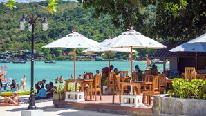 A beachside café with outdoor seating and umbrellas, overlooking a turquoise sea with boats and a forested hill in the background.