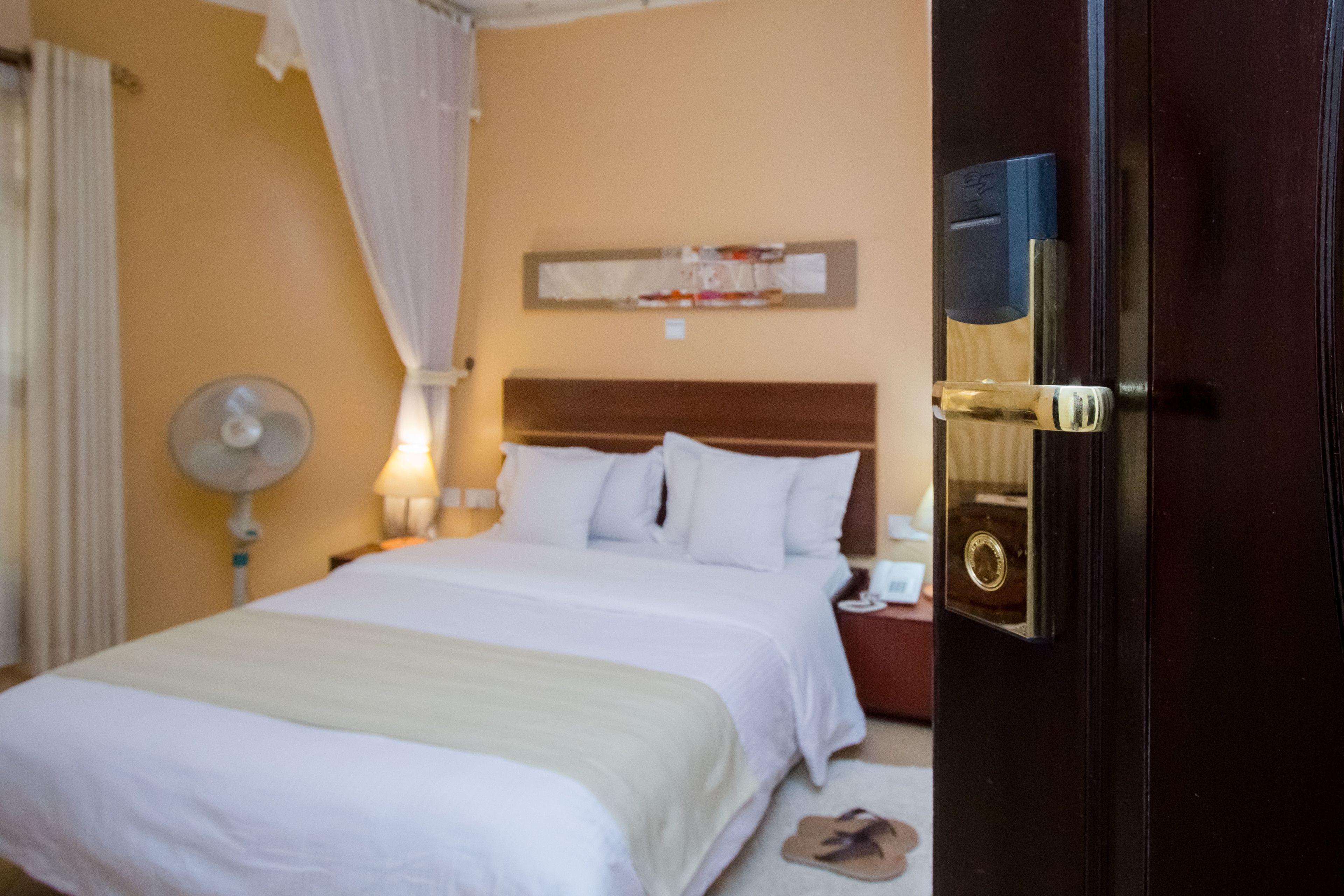 Hotellihuone, jossa on suuri sänky, yöpöytä lampun kanssa, seinälle ripustettu taideteos ja ovi, jossa on avaimenreikä.