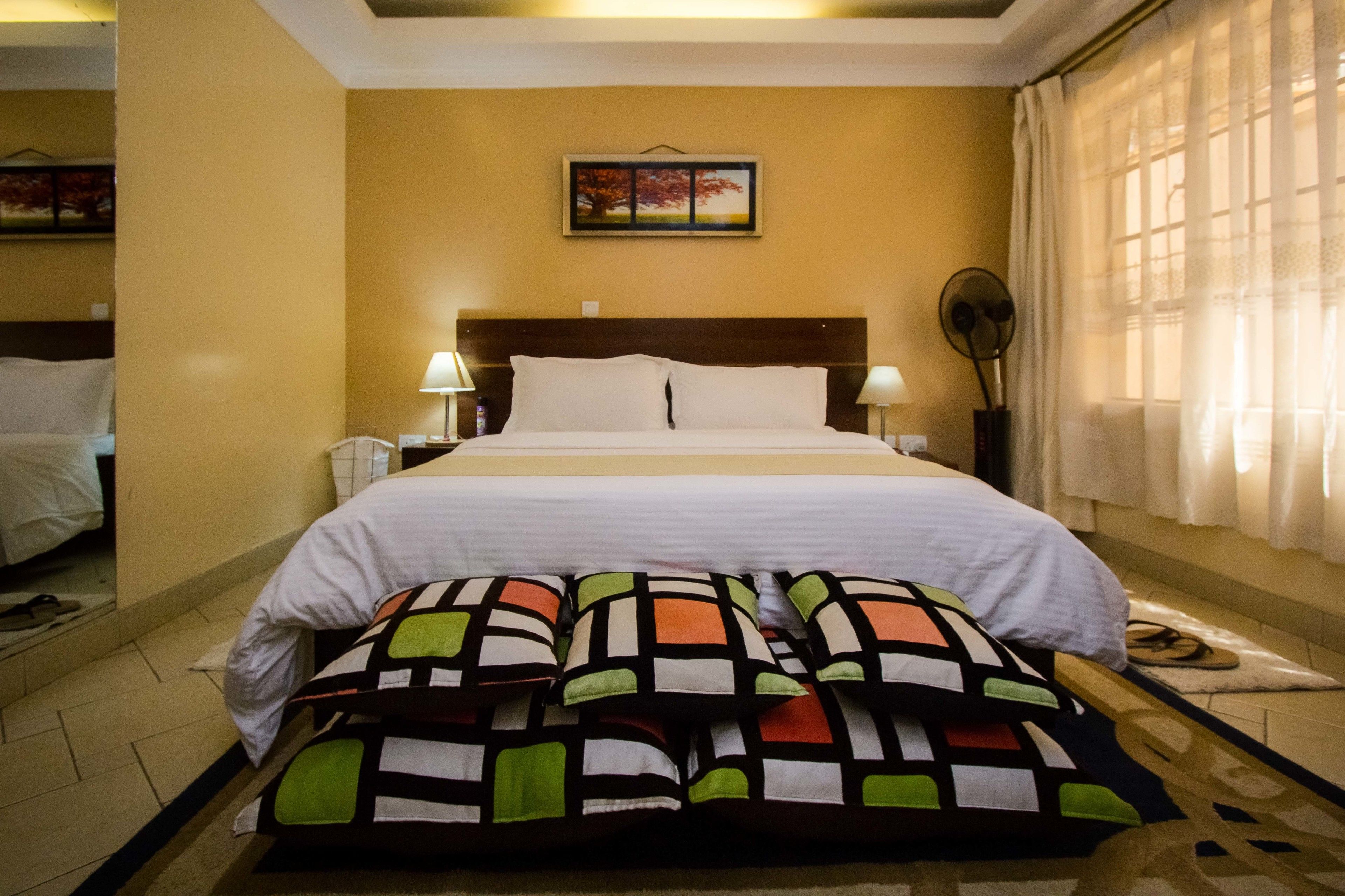 A hotel room with a bed, pillows, a nightstand, a lamp, a mirror, and a window with curtains.