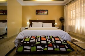 A hotel room with a bed, pillows, a nightstand, a lamp, a mirror, and a window with curtains. - Capital Heights Hotel (Nairobi)