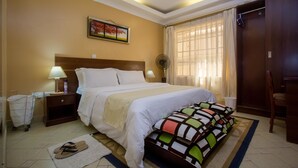Habitación de hotel con una cama grande, una mesita de noche con lámpara, un ventilador, una ventana con cortinas y una puerta.