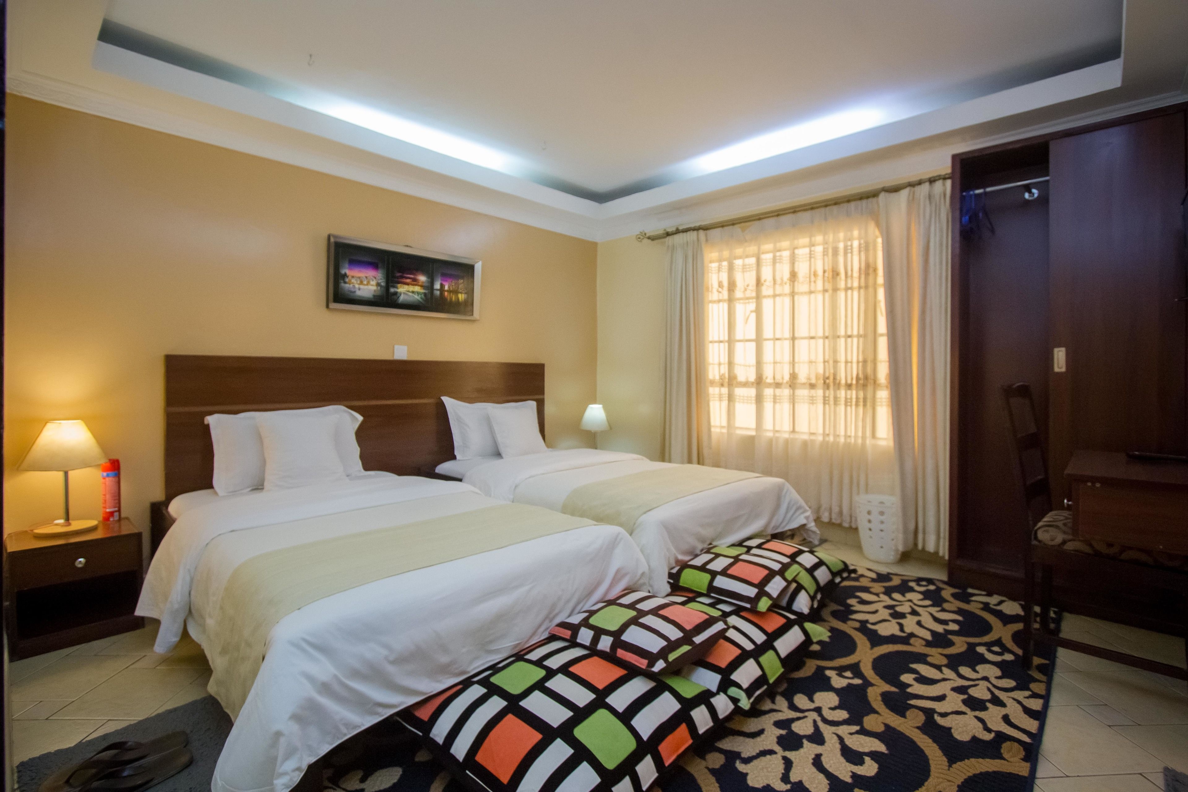 A hotel room with two beds, a nightstand, a lamp, a window with curtains, and a door.