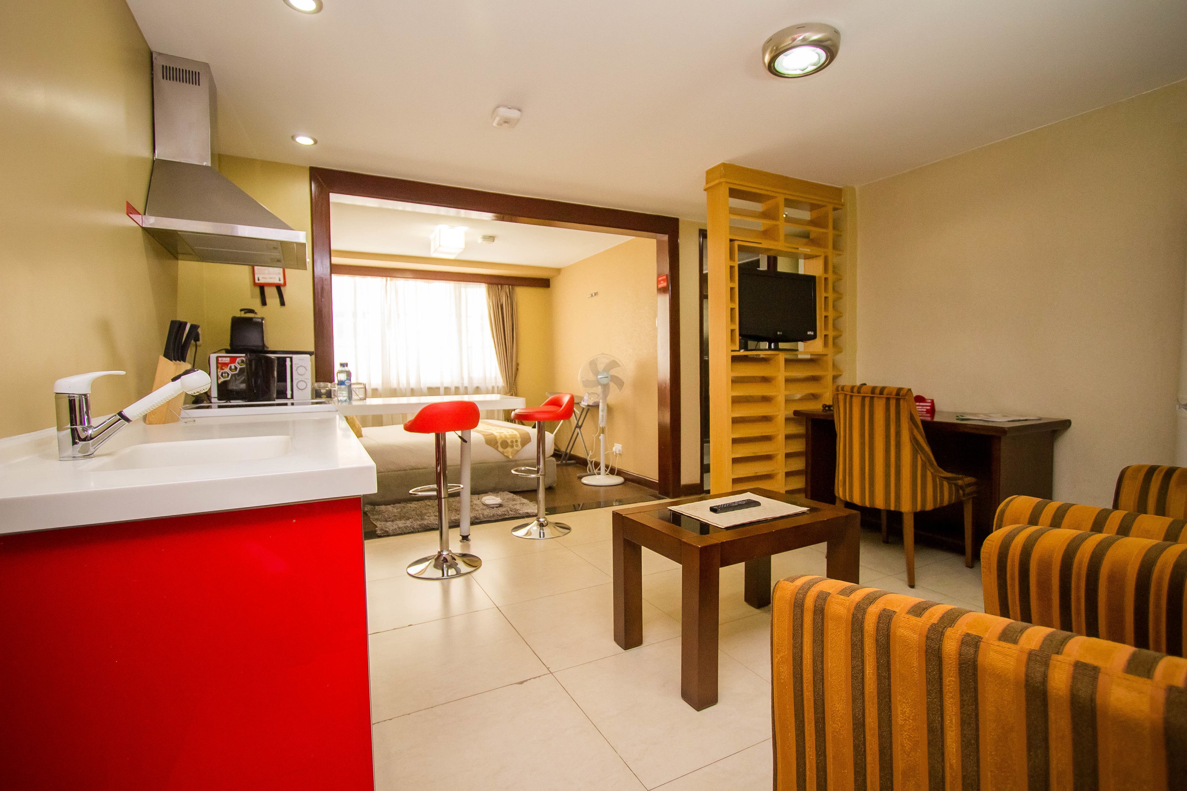 Habitación de hotel con kitchenette, comedor con mesa y sillas, televisor montado en un panel de madera y un amplio ventanal con cortinas.