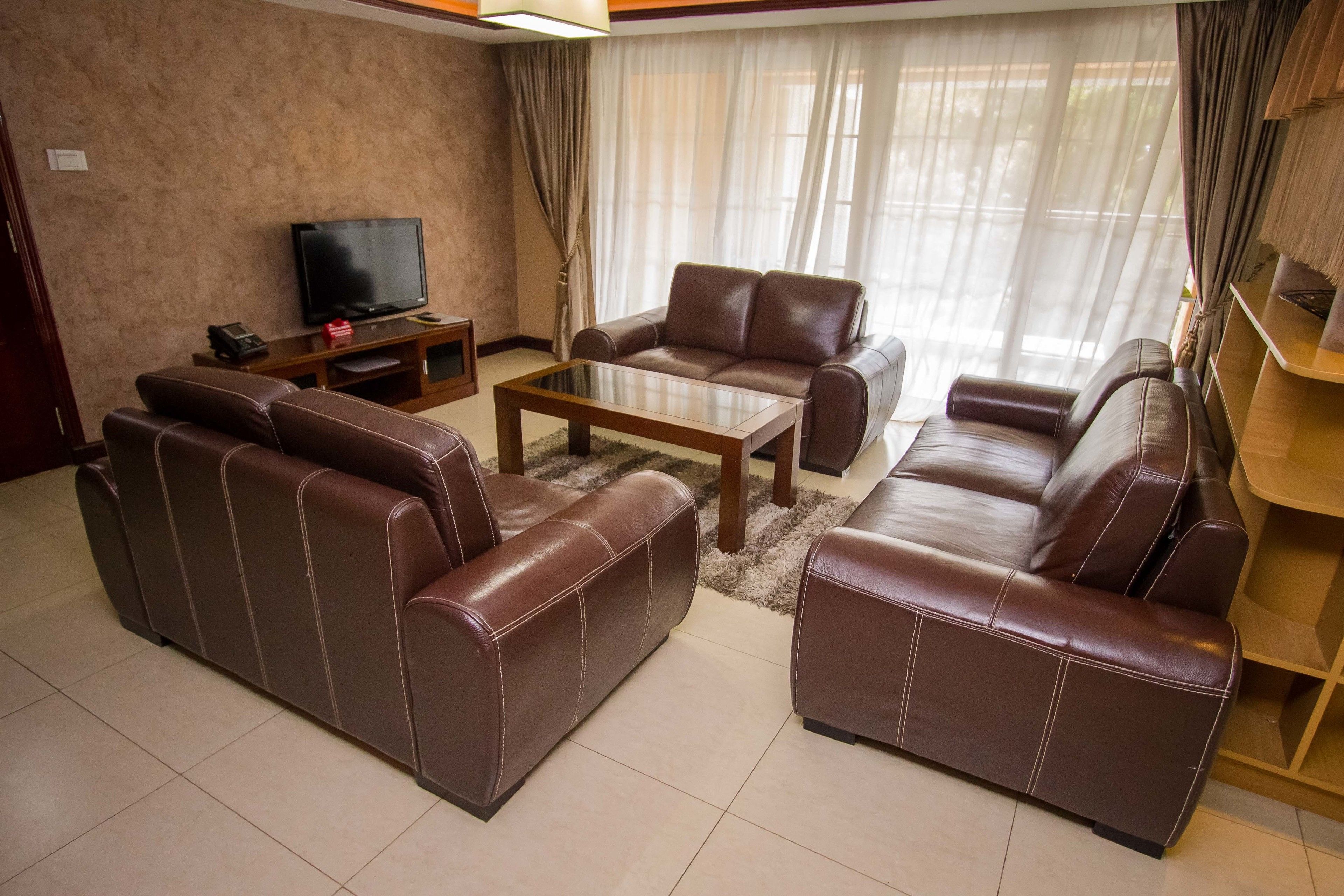 A living room with a brown leather sofa set, a glass coffee table, a flat-screen TV on a wooden stand, and a bookshelf.