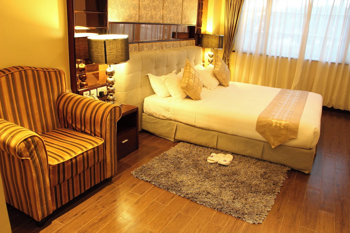 A hotel room with a bed, a striped armchair, a nightstand with a lamp, and a pair of slippers on the rug.
