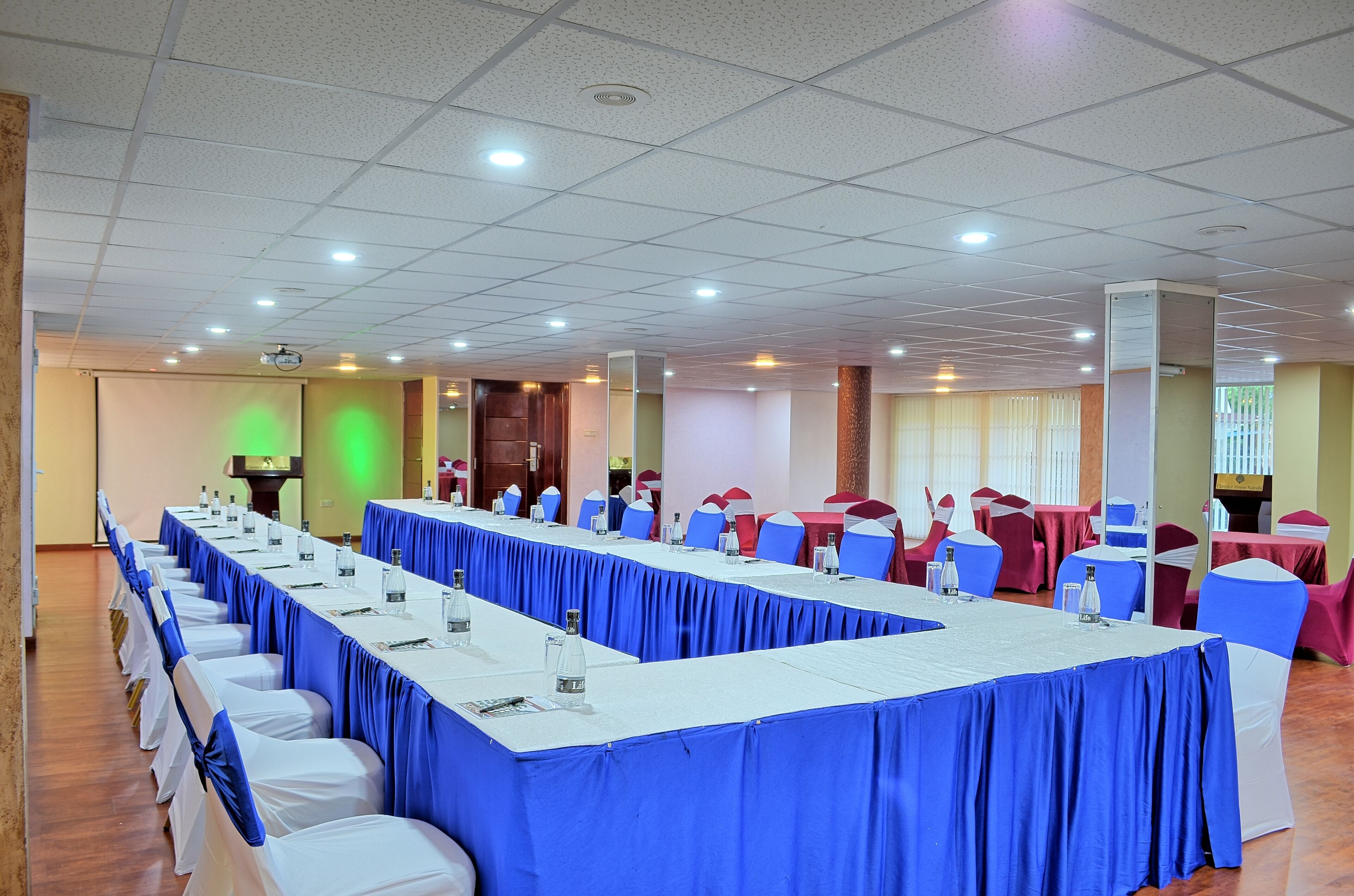 A conference room with long tables, blue and white chairs, and a projector screen.
