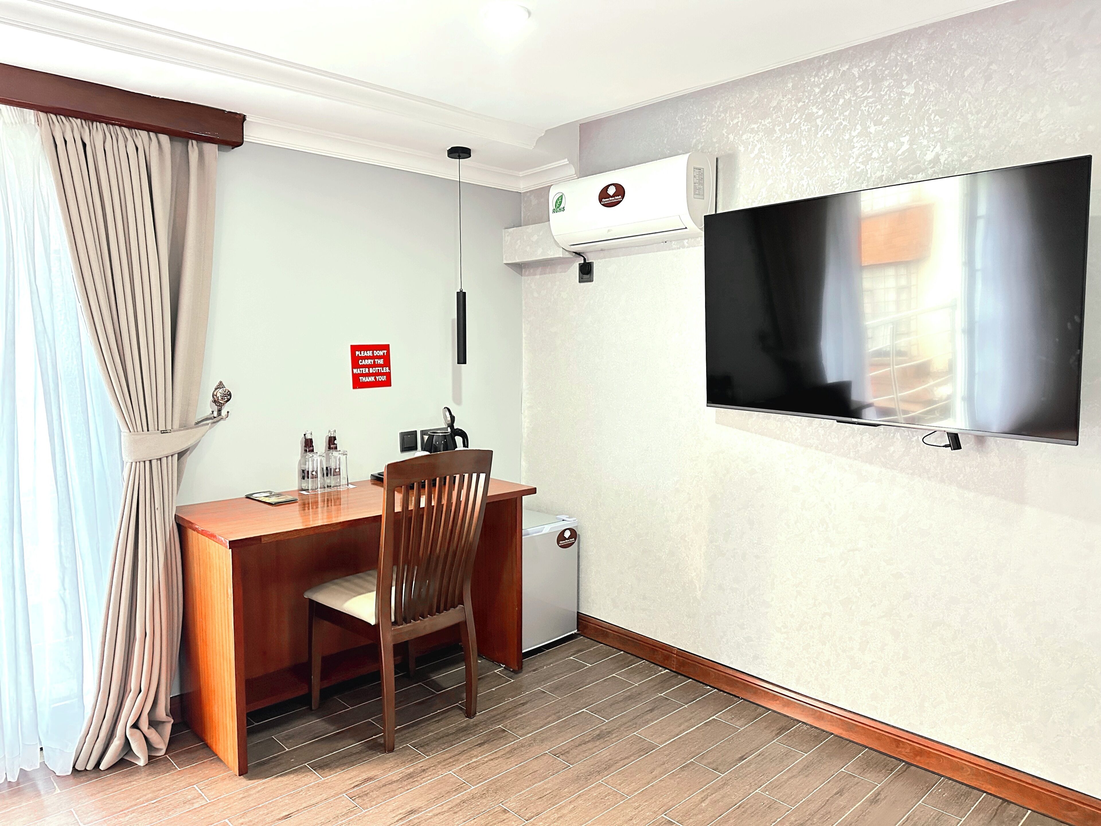 Een hotelkamer met een houten bureau, een stoel, een aan de muur bevestigde televisie, een airconditioning en een rode bordje aan de muur.