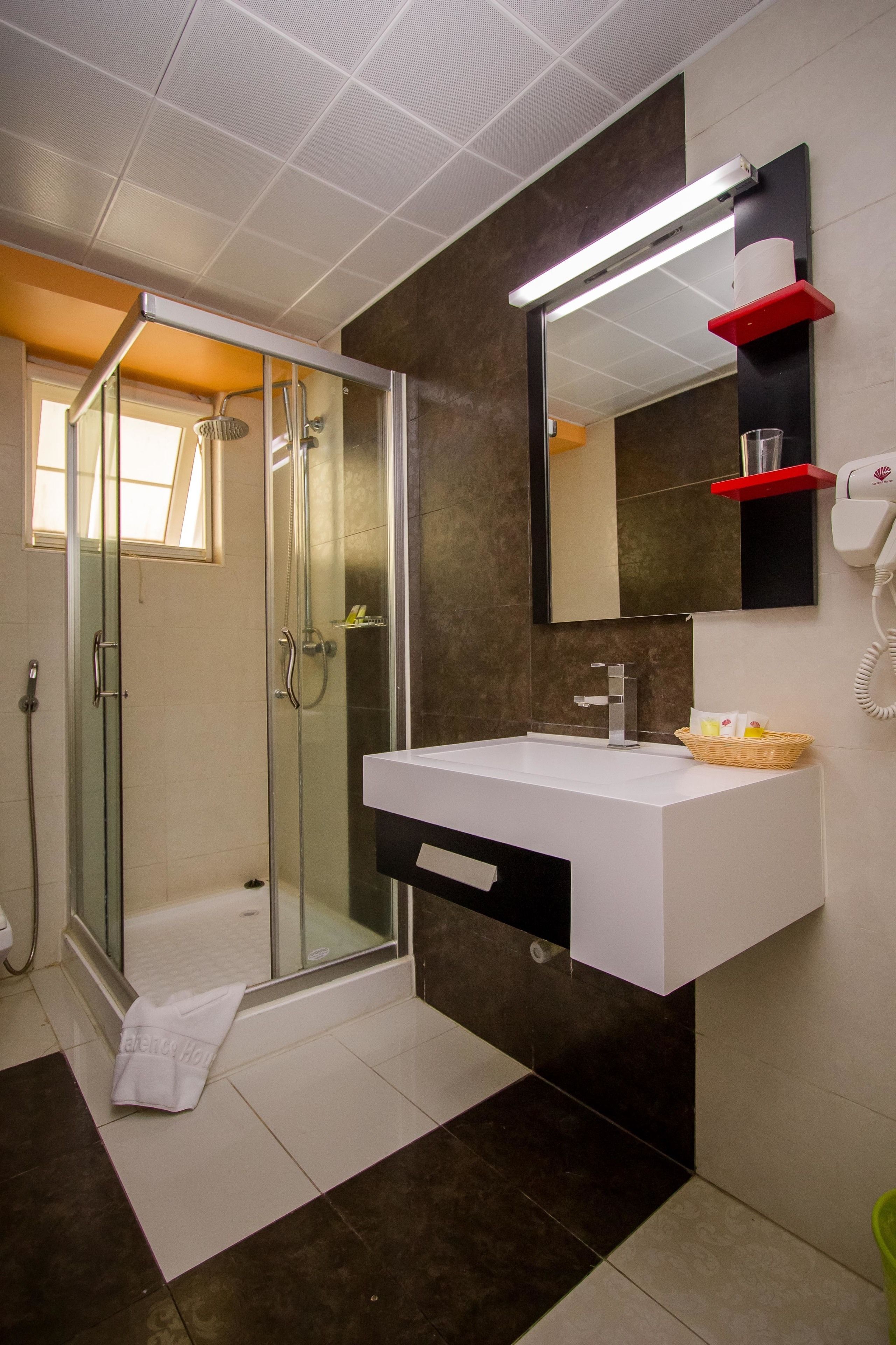 Uma casa de banho moderna com cabine de duche em vidro, lavatório branco com bancada preta e espelho com prateleira vermelha.