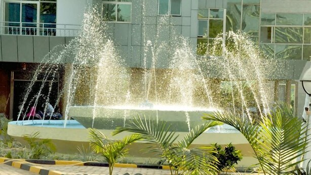 Une fontaine dotée de plusieurs jets d’eau, située dans un espace paysager agrémenté de palmiers, avec un bâtiment en arrière-plan.