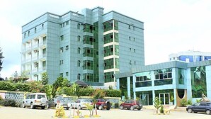 Un immeuble à plusieurs étages doté d’une façade moderne en verre, un parking avec plusieurs voitures et un petit jardin agrémenté de plantes tropicales.