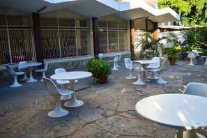 An outdoor patio area with white tables and chairs, stone flooring, and potted plants.