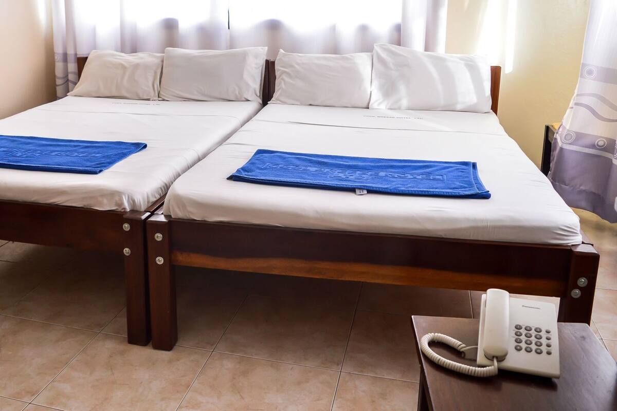 Een tweepersoonsbed met witte lakens en blauwe handdoeken, een telefoon naast het bed, en een raam met gordijnen.