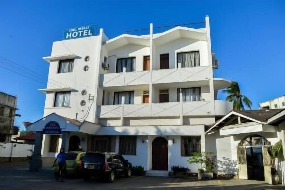 Een meerlaags hotel met een bord dat aangeeft dat het een hotel is. Er staan auto's geparkeerd voor en een persoon loopt naar de ingang.
