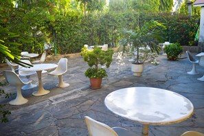 Eine Terrassenfläche mit Steinböden, weißen Tischen und Stühlen sowie verschiedenen Kübelpflanzen.