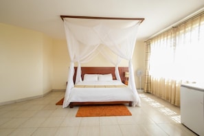 Una cama con dosel con ropa de cama blanca y un tapete marrón en una habitación de hotel, con cabecera de madera, un ventilador de pie y una ventana con cortinas.