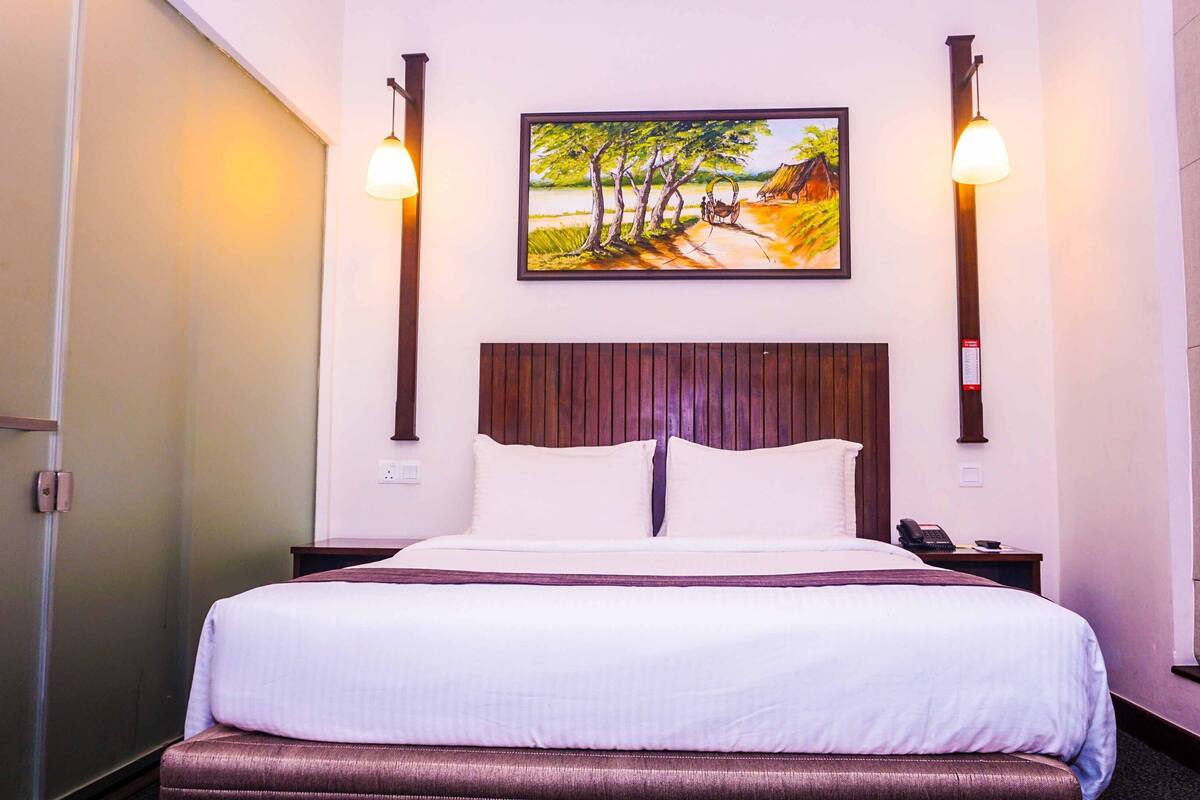 A hotel room with a wooden headboard, a painting of a rural scene, a bedside lamp, and a telephone on the nightstand.