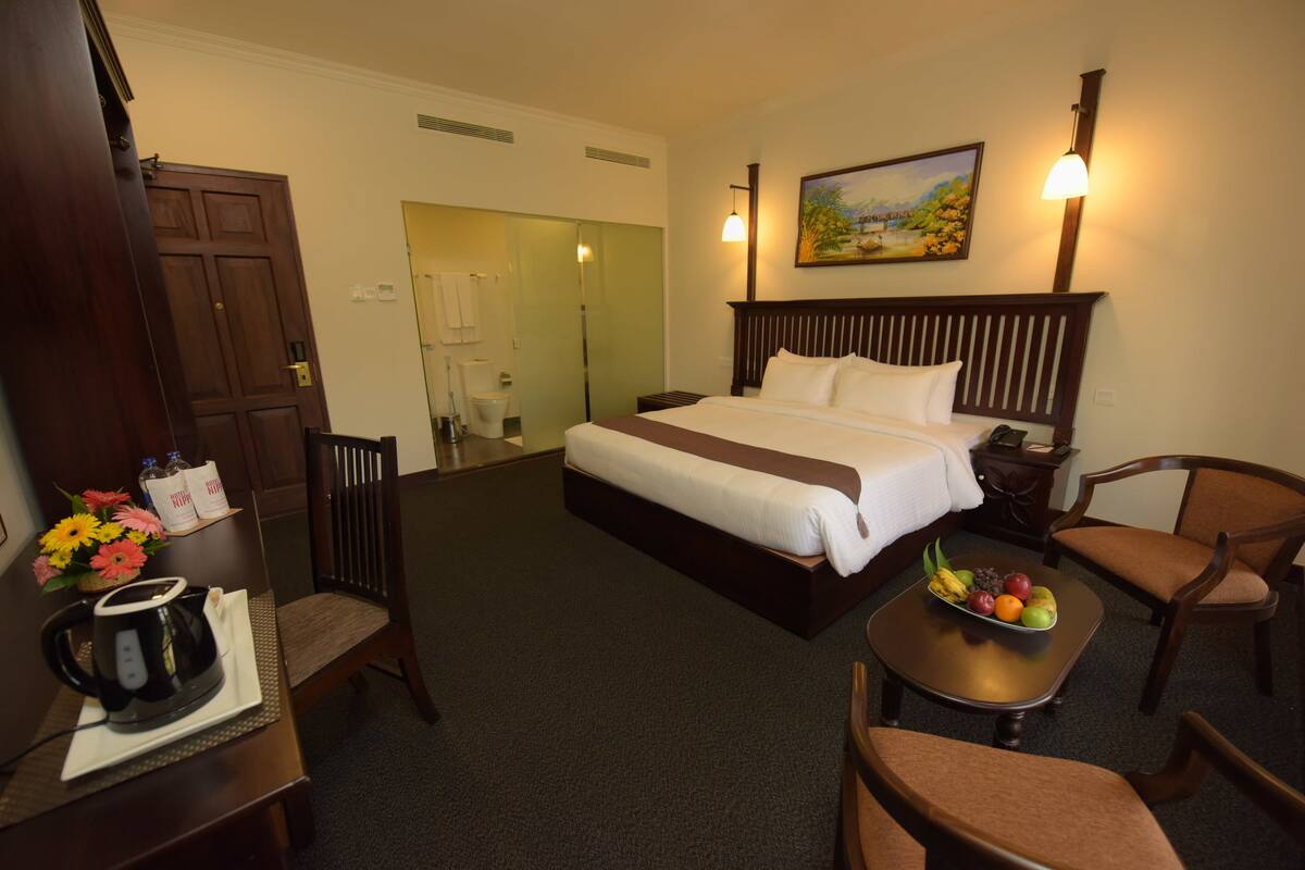 A hotel room with a large bed, a desk with a kettle, a fruit bowl, and a painting on the wall.