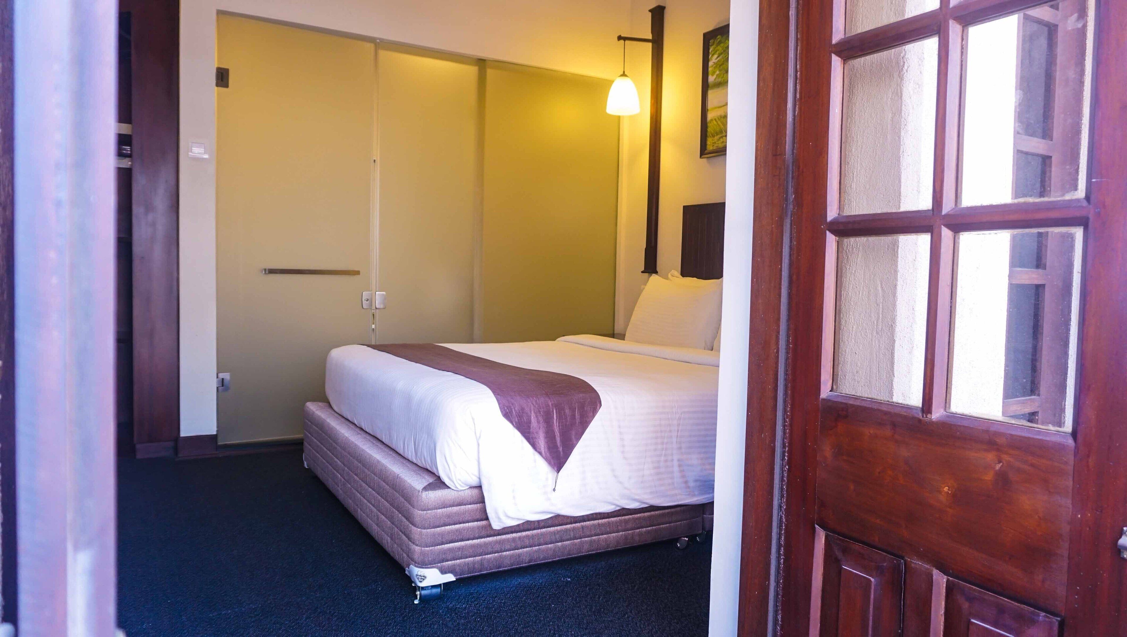 A hotel room with a bed, a bedside table, a lamp, and a door.