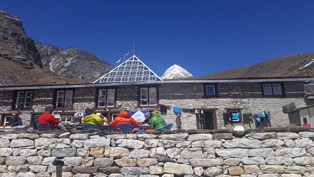 A stone building with a triangular roof, people sitting outside, and a mountain in the background. - 8000 Inn (Lobuche)