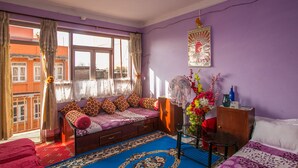 Quarto com cama, janela com cortinas, ventilador, vaso com flores e pôster na parede.