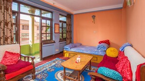 Quarto com sofá, mesa de madeira, cama com roupa de cama colorida, tapete estampado e janelas amplas com cortinas.