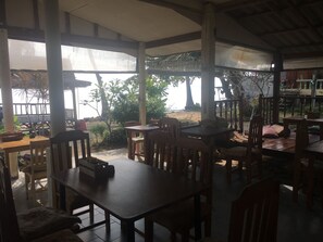 A covered outdoor dining area with wooden tables and chairs, surrounded by plants and a view of the beach.