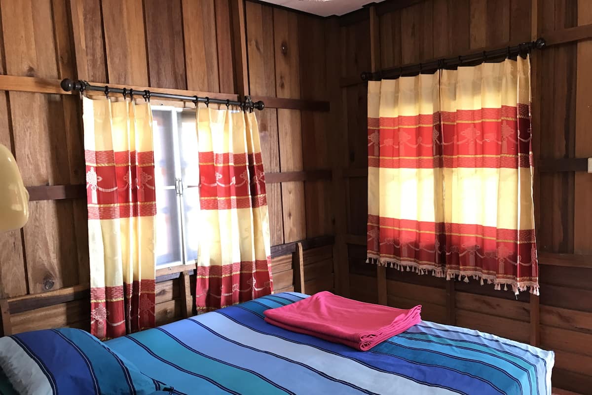 Una habitación de cabaña de madera con una cama, dos ventanas con cortinas y un cabecero de madera.