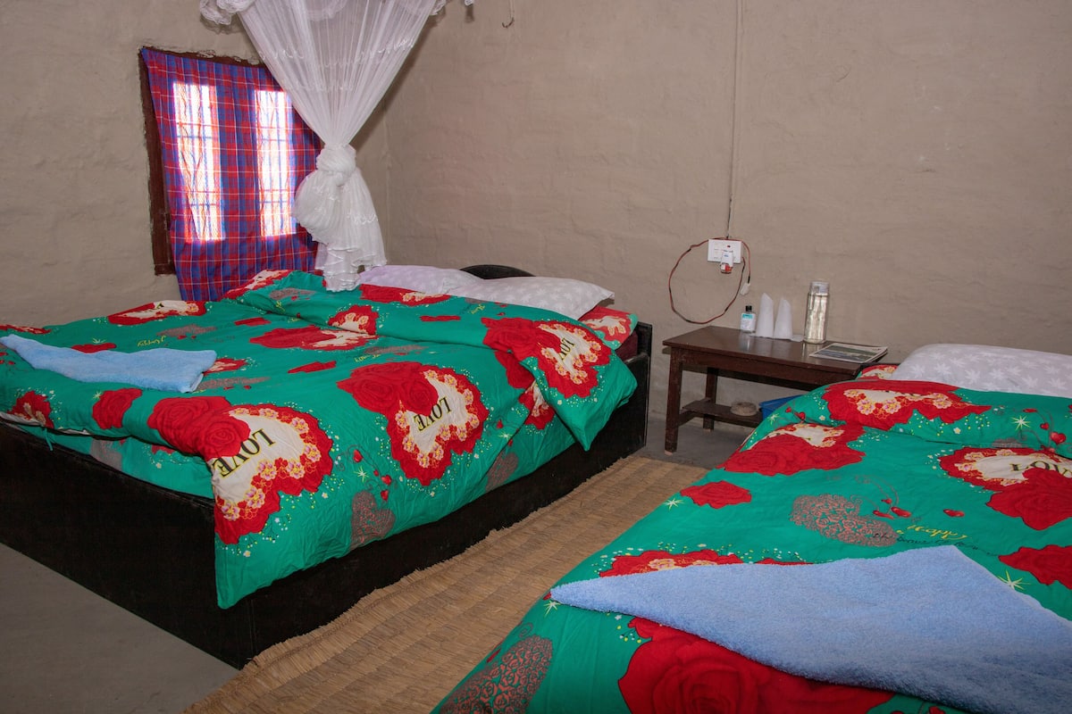 A room with two beds, a bedside table, a window with a curtain, and a wall socket.