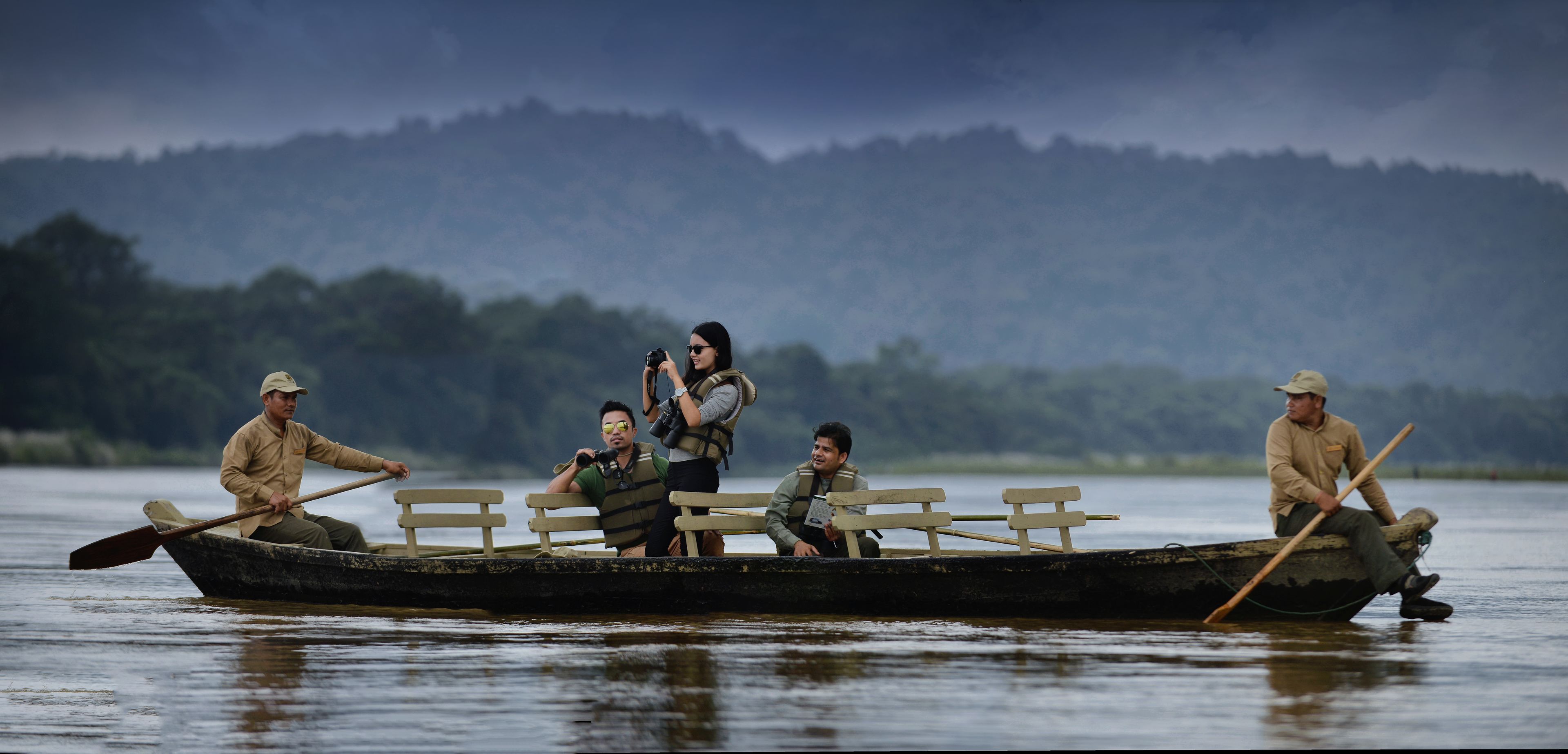 A rowboat with four people, one taking a photo, on a calm body of water with mountains in the background.
