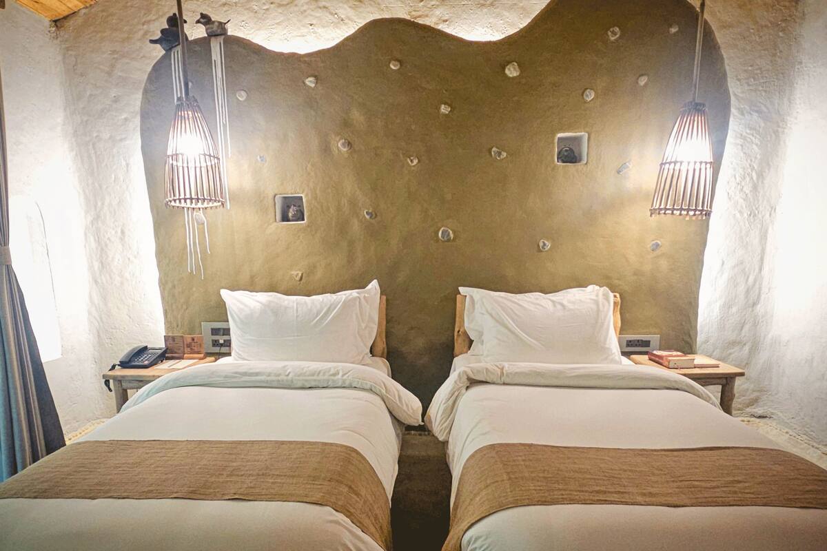 A room with two beds, each with white linens and a brown headboard. There are two bedside tables with lamps and a small picture frame on each. The walls are textured and have a rustic appearance.
