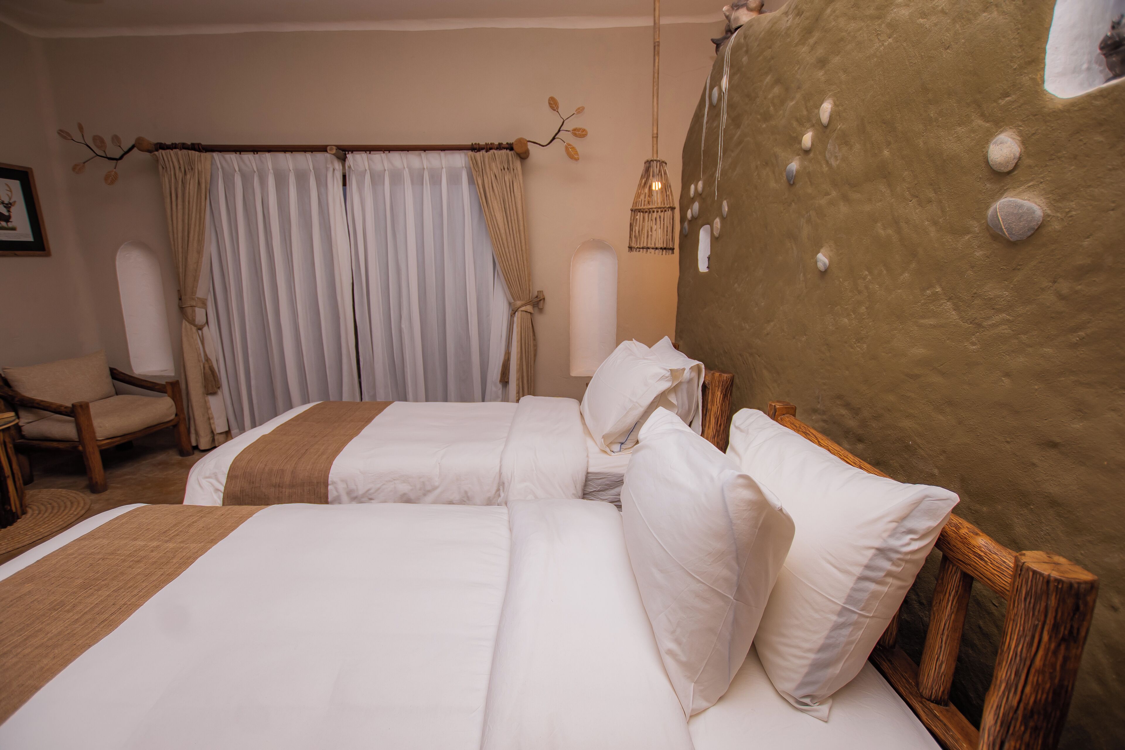 Quarto de hotel com duas camas, uma cadeira, uma janela grande com cortinas e uma parede com textura diferenciada.