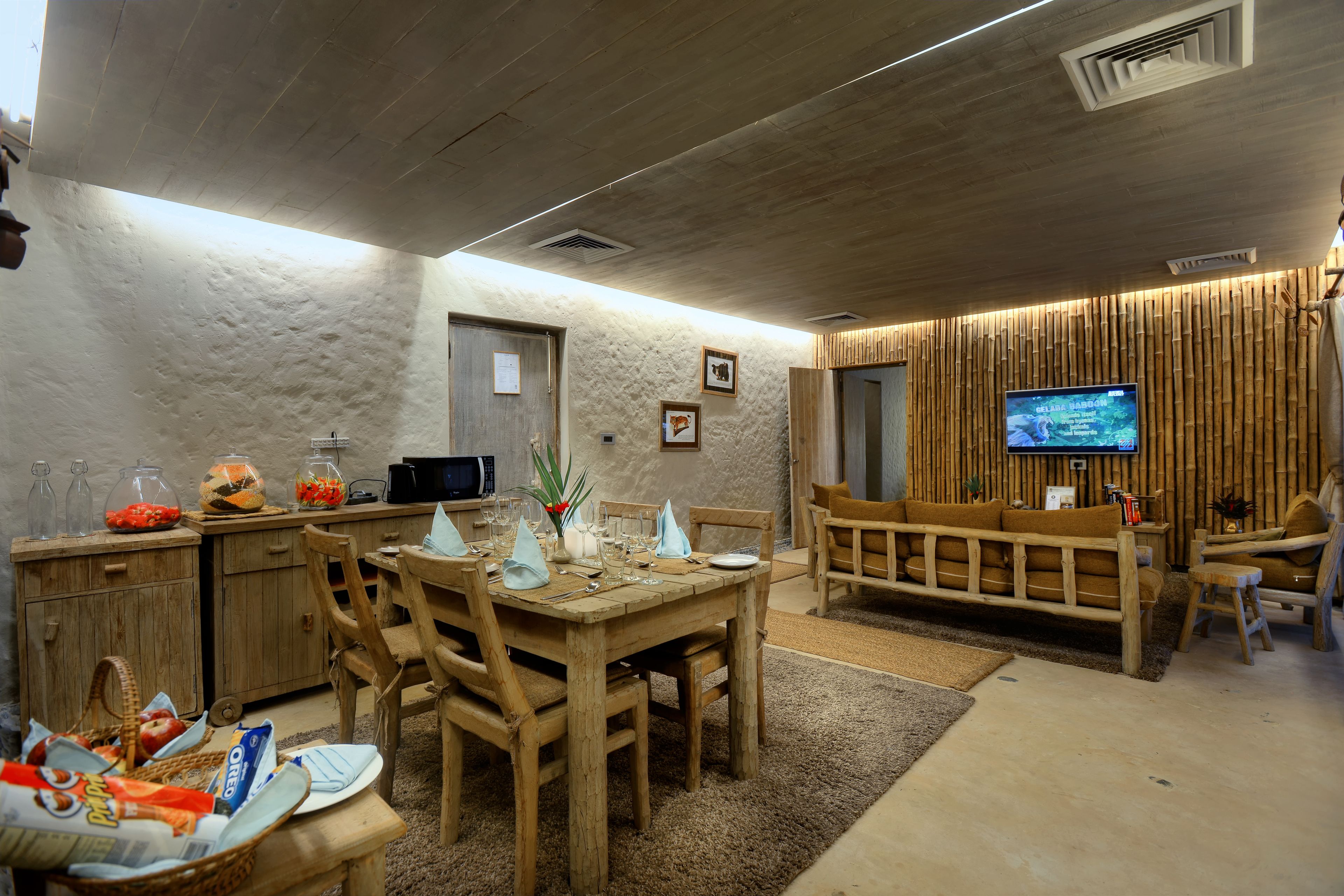 A dining area with a wooden table set for four, a television mounted on a bamboo wall, and a kitchen area with a microwave and a refrigerator.