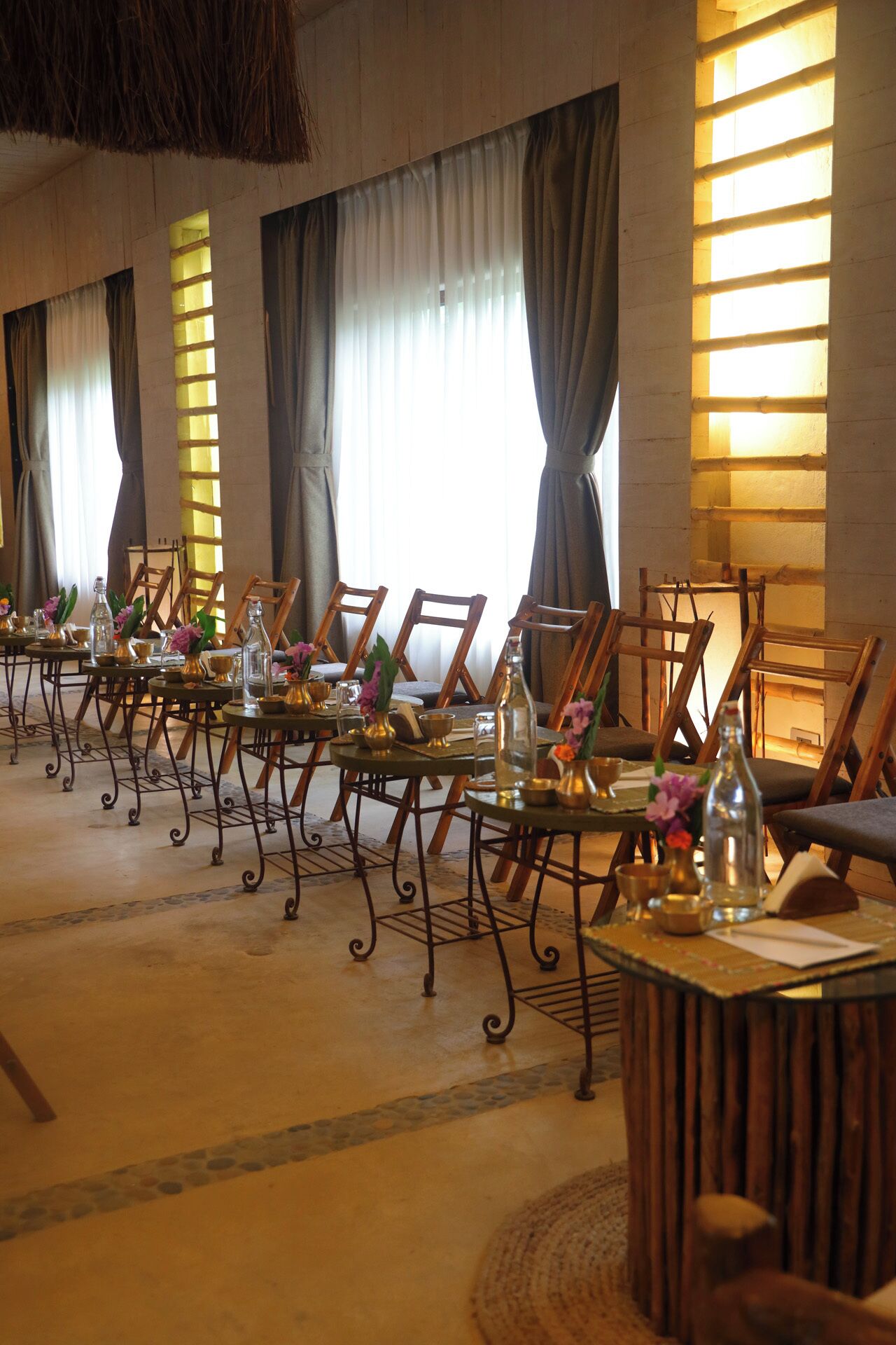 A long dining table with chairs, set with glassware and flowers, in a room with large windows and curtains.