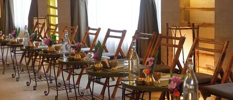A long dining table with chairs, set with glassware and flowers, in a room with large windows and curtains.