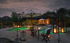Un área de comedor al aire libre con piscina, suelo de piedra y construcciones con techo de paja.