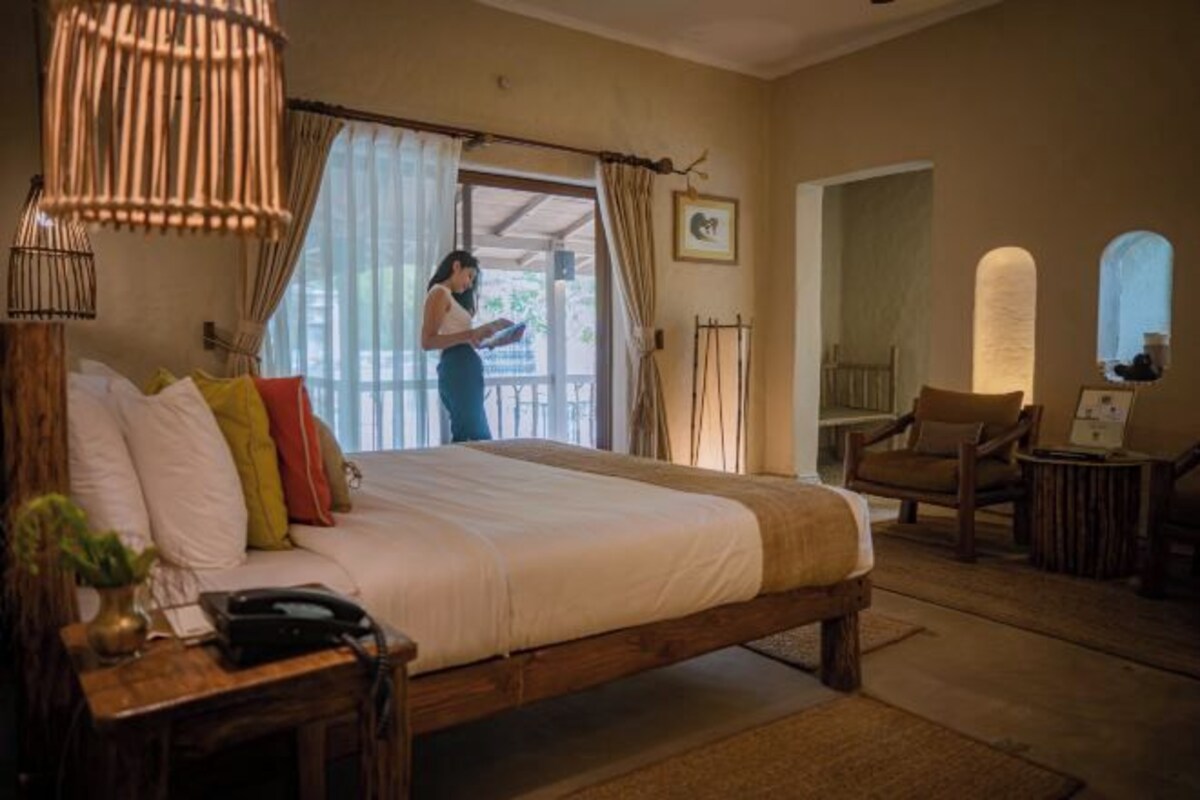 Un dormitorio con cama de madera, una mesita de noche con un teléfono, una lámpara y una silla. Hay una ventana con cortinas y vistas al exterior.