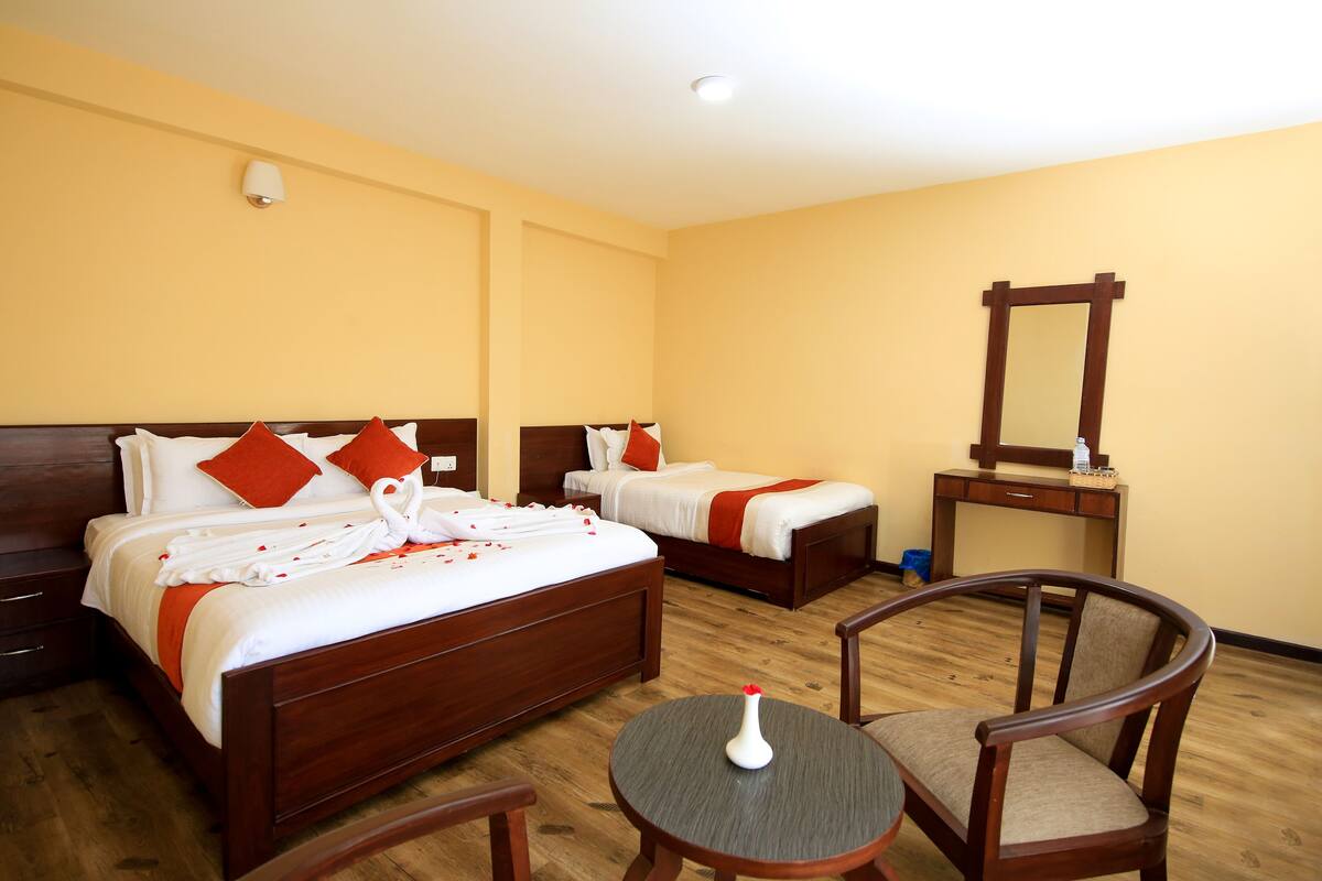 A hotel room with two wooden beds, each with white bedding and orange pillows, a round table with a vase, and a mirror on a wooden stand.