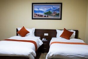 Habitación de hotel con dos camas, cada una con ropa de cama blanca y detalles naranja, una mesita de noche con teléfono y cargador, y un cuadro en la pared con un paisaje montañoso.