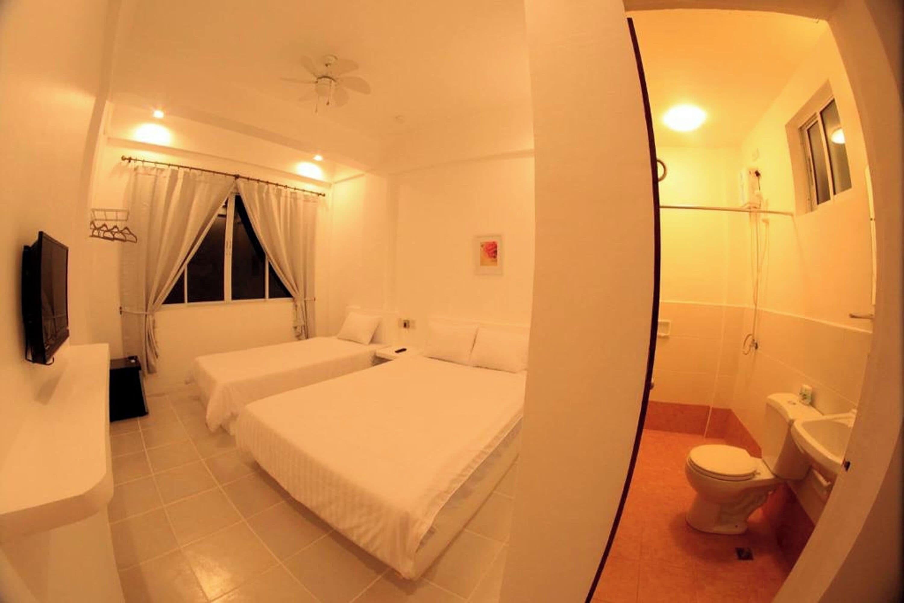 A room with two beds, a ceiling fan, a window with curtains, a small table, and a bathroom with a toilet and sink.