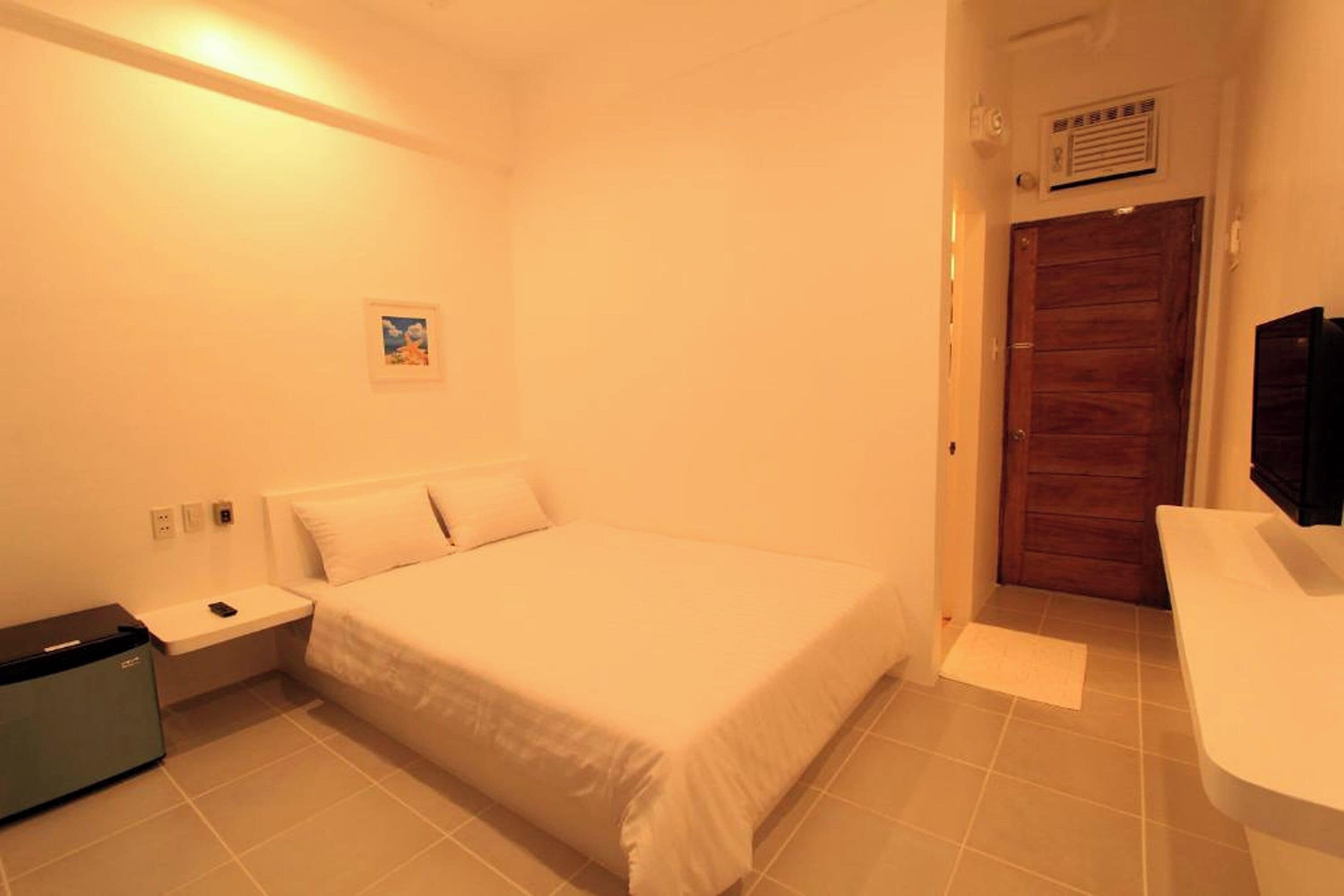Un dormitorio con cama, mesita de noche, una pequeña mesa, un calentador en la pared y una puerta.