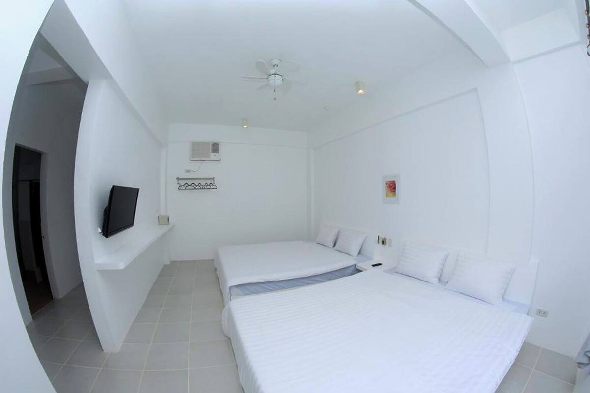 Un dormitorio con dos camas, un ventilador de techo, un televisor en un estante y un pequeño adorno en la pared.