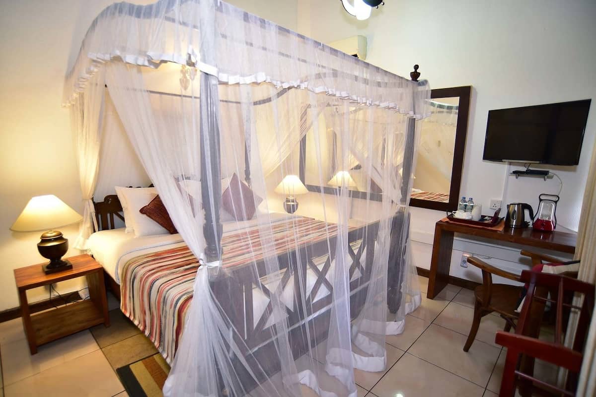 A hotel room with a four-poster bed, a desk with a chair, a television, and a bedside table with a lamp.