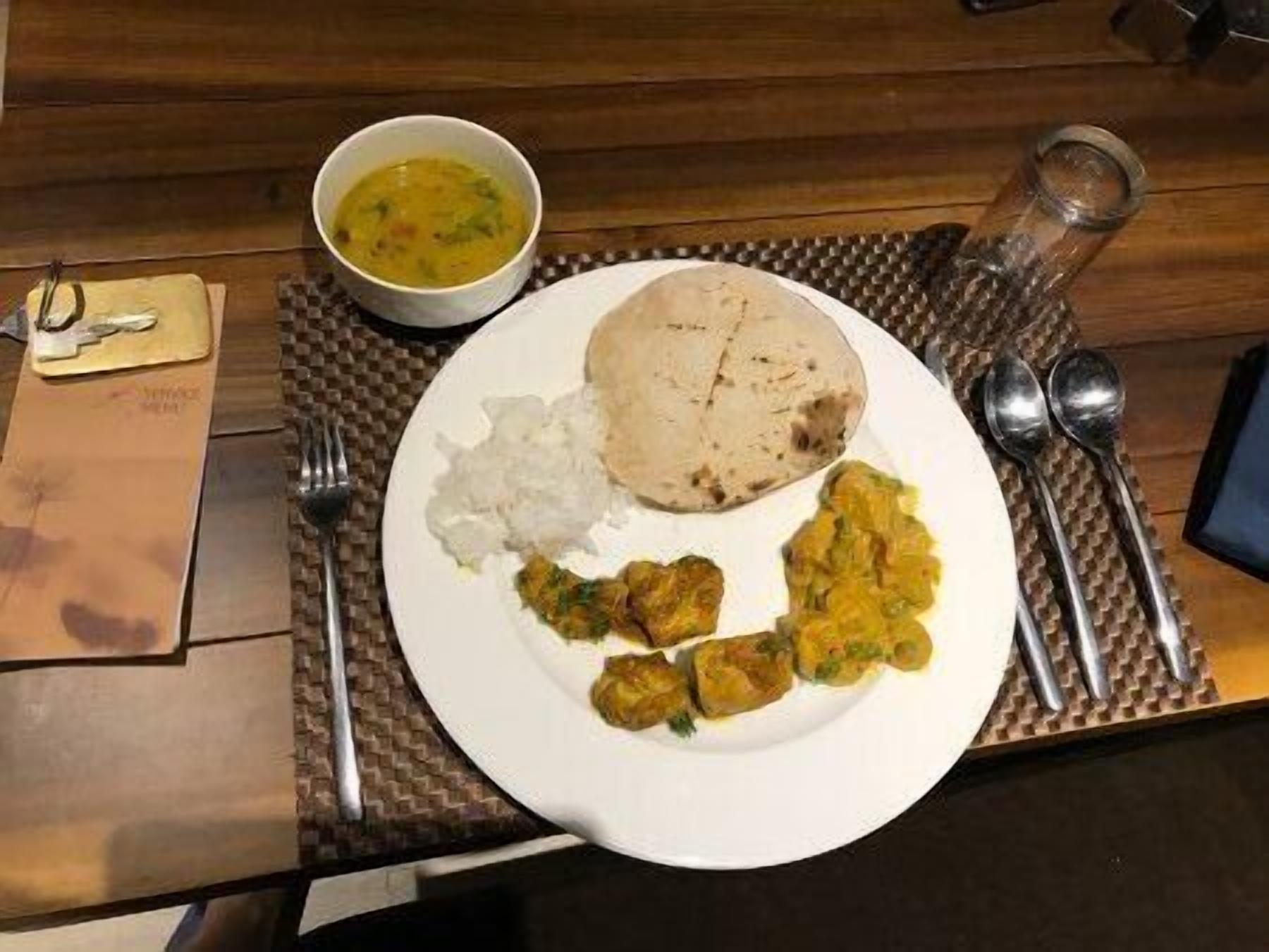A plate with rice, a flatbread, and curry, accompanied by a bowl of soup, on a wooden table with a placemat.